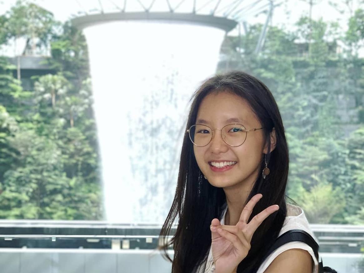 woman with black hair wearing glasses and smiling with her hands in the peace symbol is standing in front of a fountain and green trees